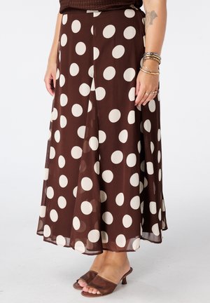 Jupe midi marron à gros pois blancs portée avec des sandales marron à talons ouverts et des bracelets au poignet droit.