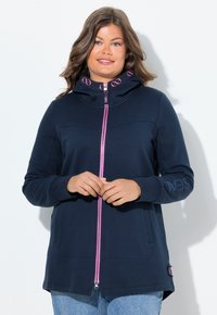 Donna che indossa una felpa con zip blu navy con zip e dettagli del logo rosa, in piedi contro uno sfondo bianco con le mani delicatamente incrociate.