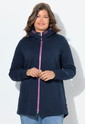 Donna che indossa una felpa con zip blu navy con zip e dettagli del logo rosa, in piedi contro uno sfondo bianco con le mani delicatamente incrociate.