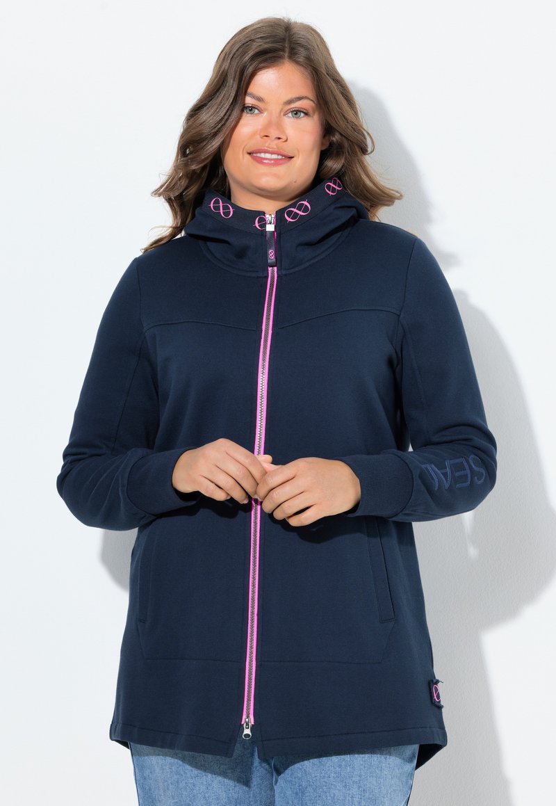 Donna che indossa una felpa con zip blu navy con zip e dettagli del logo rosa, in piedi contro uno sfondo bianco con le mani delicatamente incrociate.