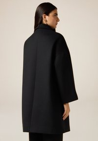 Abrigo de lana negro con cuello alto, silueta oversize, hombros estructurados y mangas de tres cuartos, mostrado desde atrás.