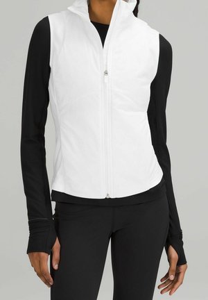 White sleeveless zip-up vest worn over a black long-sleeve top and black leggings on a standing person against plain gray background.