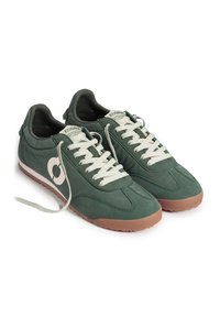 Paire de baskets basses vert foncé avec des lacets blancs, des semelles beiges et un logo circulaire blanc sur le côté. Un lacet est défait.