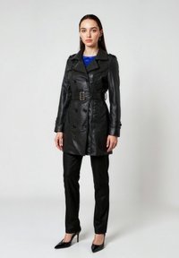 Zwarte leren trenchcoat met dubbele rij knopen, taille met ceintuur en brede kraag, gecombineerd met glanzende zwarte broek en hoge hakken.