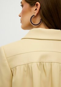 Beige maßgeschneiderte Jacke mit einem gefältelten Rückendetail, kombiniert mit großen runden Ohrringen in einem burdeos- und Goldfarbschema.