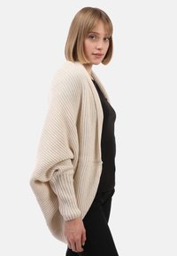 Cardigan beige oversize avec texture côtelée, manches larges et poches avant, porté sur un haut noir. L'arrière-plan est blanc uni.