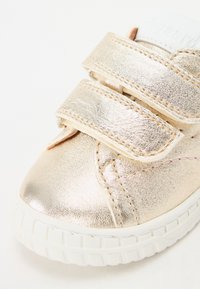 Złote metaliczne sneakersy z dwoma paskami na rzepy, białą gumową podeszwą i przeszytymi akcentami. Gładka tekstura z zaokrąglonym designem noska.