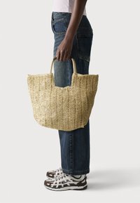 Sac en paille tressée avec un grand dessus ouvert, des poignées ergonomiques et une finition naturelle texturée. Associé à un jean bleu et des chaussures de sport.