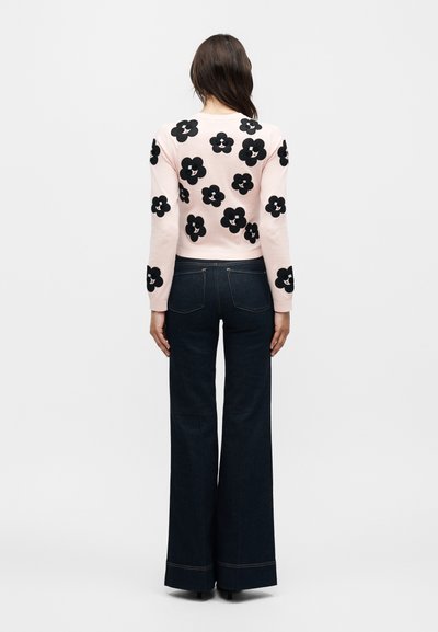 Γυναίκα που φορά ανοιχτό ροζ πουλόβερ με μαύρα λουλουδένια σχέδια και σκούρο τζιν με φαρδιά μπατζάκια, στέκεται με την πλάτη γυρισμένη σε λευκό φόντο.
