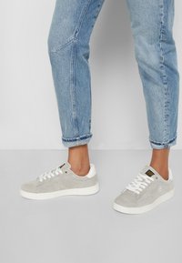 Jasnoszare zamszowe sneakersy z białymi gumowymi podeszwami i sznurowadłami, charakteryzujące się gładką teksturą i minimalistycznym designem. Widoczne są dżinsy z podwiniętymi nogawkami.