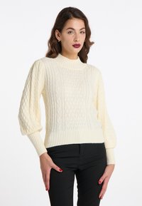 Pulover crem cu tricotaj tip cablu, cu guler înalt cu nervuri și mâneci bufante; prezintă modele texturate și un tiv ajustat. Purtați-l cu pantaloni negri.