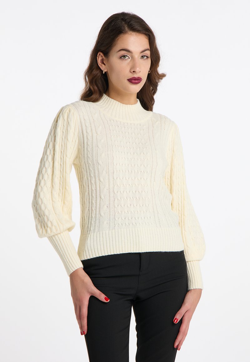 Pulover crem cu tricotaj tip cablu, cu guler înalt cu nervuri și mâneci bufante; prezintă modele texturate și un tiv ajustat. Purtați-l cu pantaloni negri.