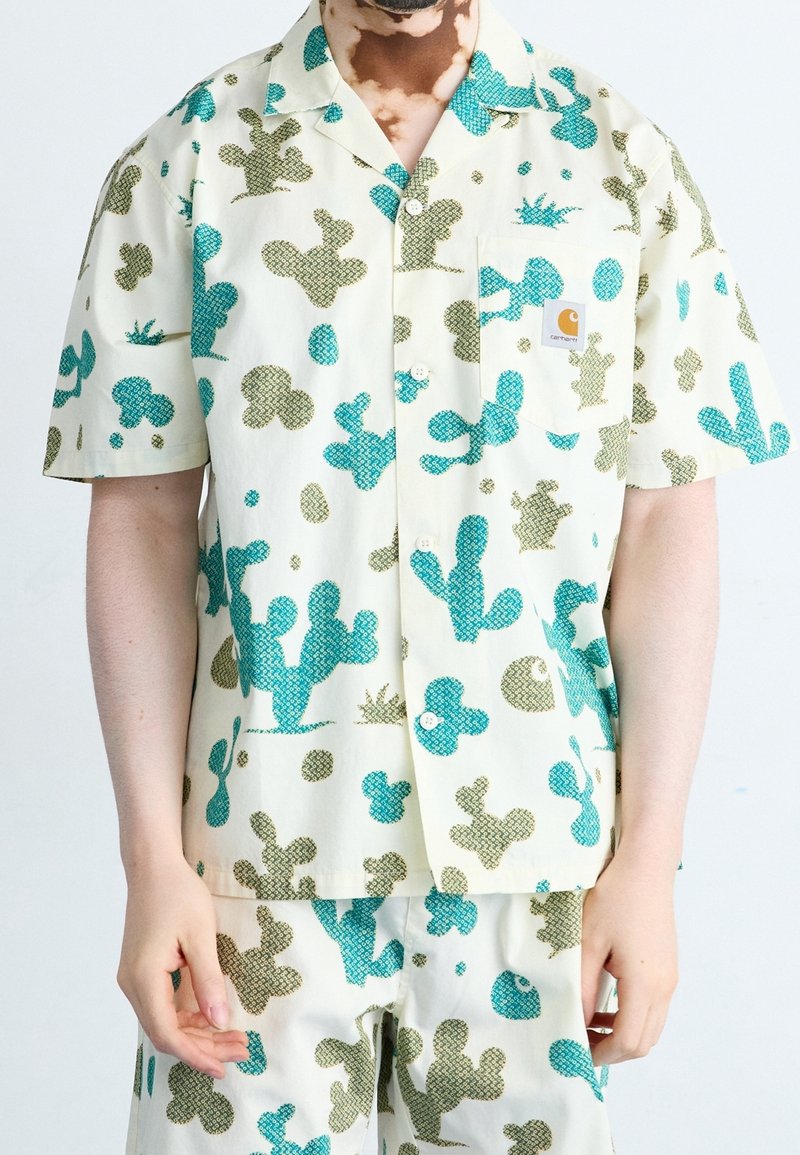 Homme portant une chemise à manches courtes boutonnée et un short assorti avec un imprimé abstrait de cactus vert et bleu sur fond crème.