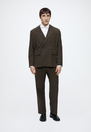Homme aux cheveux courts et foncés, portant un costume croisé marron foncé, une chemise blanche et des chaussures noires, debout devant un fond blanc.