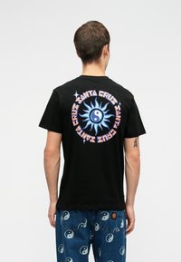 Isik, kellel on seljas must T-särk, millel on sinine päike ja yin-yangi disain ning "Santa Cruz" tekstring tagaküljel, koos siniste yin-yangi printidega teksadega.