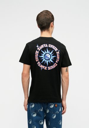 Άτομο που φοράει μια μαύρη μπλούζα με σχέδιο μπλε ήλιου και γιν-γιανγκ, και με την εγγραφή "Santa Cruz" σε κύκλο στο πίσω μέρος, συνδυασμένη με τζιν με μπλε σχέδιο γιν-γιανγκ.