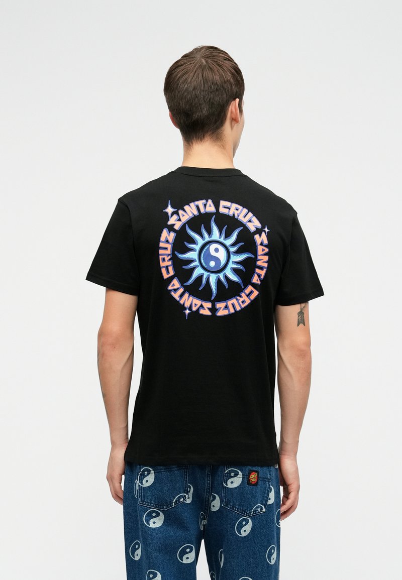 Isik, kellel on seljas must T-särk, millel on sinine päike ja yin-yangi disain ning "Santa Cruz" tekstring tagaküljel, koos siniste yin-yangi printidega teksadega.