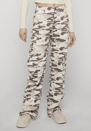 Personne portant un pantalon cargo camouflage beige, marron et blanc ample avec poches latérales et des baskets beige, debout devant un fond uni.