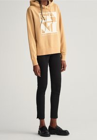 Sweat à capuche beige avec logo graphique blanc, pantalon ajusté noir et mocassins noirs. Le sweat à capuche est doté d'un cordon de serrage et de poignets côtelés.