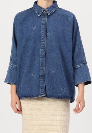 Giubbotto di denim blu scuro con chiusura a bottoni, vestibilità oversize e trafori a forma di cuore. Presenta un colletto classico e maniche larghe.