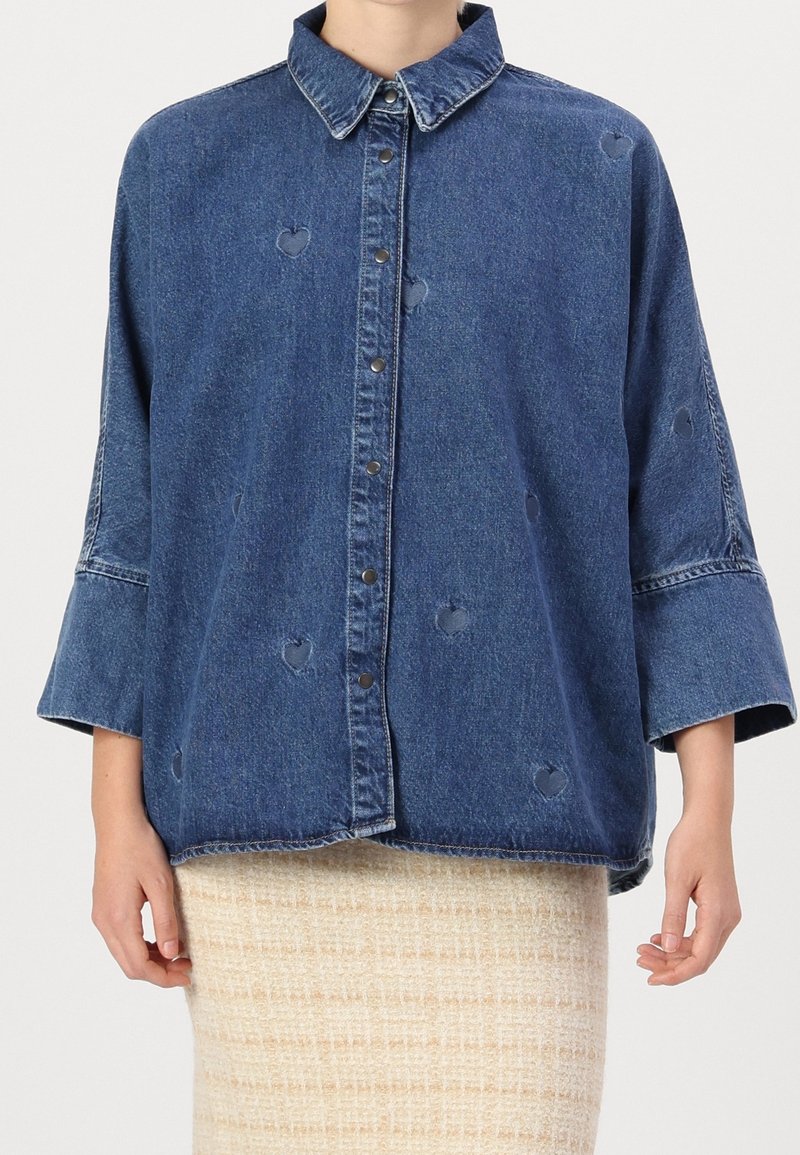 Giubbotto di denim blu scuro con chiusura a bottoni, vestibilità oversize e trafori a forma di cuore. Presenta un colletto classico e maniche larghe.