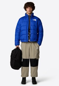 Giacca a vento blu con chiusura zip, pantaloni cargo a blocchi di colore marrone e nero, e zaino nero. Capelli ricci e stivali neri.