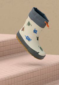 Bottes de pluie pour enfants avec des imprimés de monstres colorés en cartoon, une bordure grise, un cordon orange et une semelle jaune texturée, placées sur des marches carrelées roses.