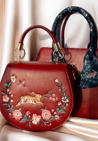 Rød lædertaske med blomsterbroderi og design af en kanin, med guldfarvet hardware og et mønstret håndtag på en sekundær taske.