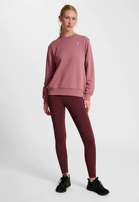 Sweat-shirt rose clair avec encolure ronde et ourlet côtelé, associé à des leggings bordeaux. Le modèle porte des chaussures de sport noires.