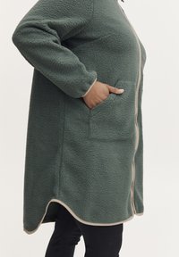 Abrigo de forro polar verde con una superficie texturizada, que cuenta con bolsillos laterales y un ribete beige a lo largo del zipper y el dobladillo, con un fondo redondeado.