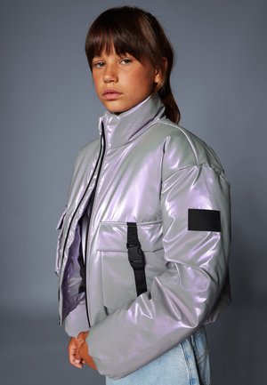 Wypełniona, metaliczna lawendowa kurtka bombowiec z wysokim kołnierzem, czarnymi detalami na paskach i naszywką na lewym ramieniu. Stylizowana z jasnoniebieskimi dżinsami.