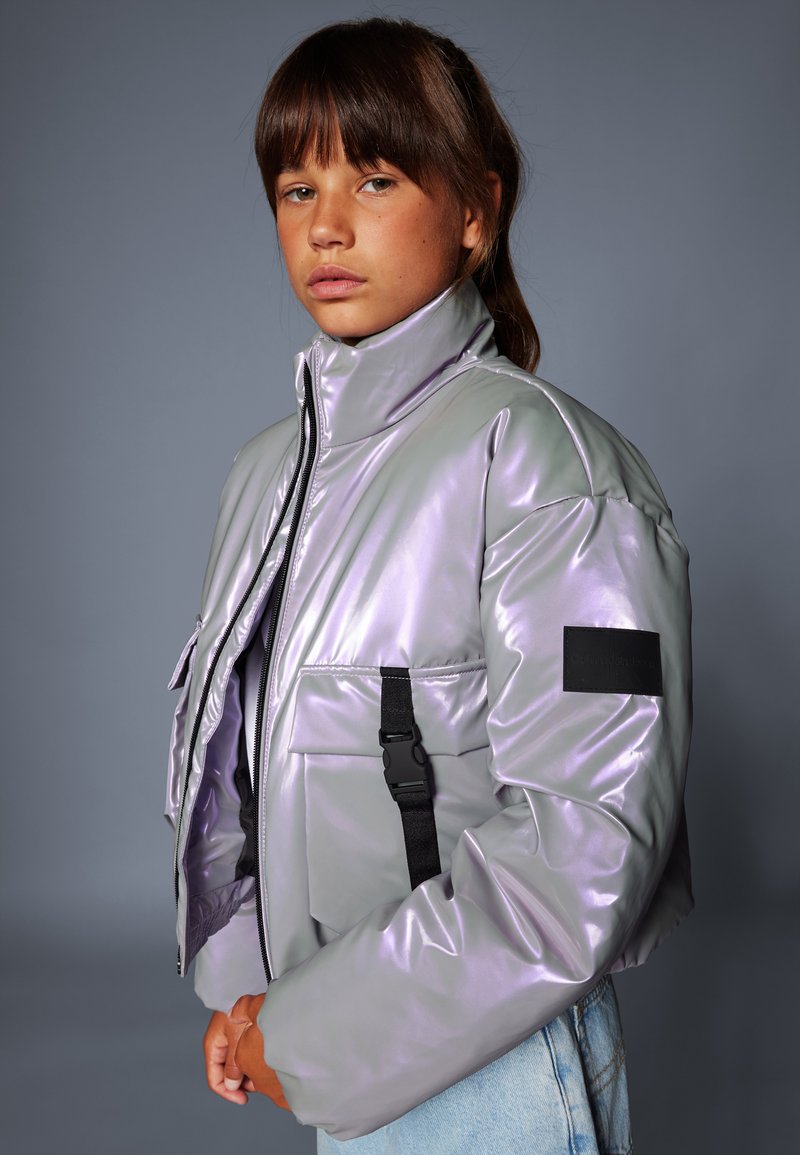 Βαμβακερή μεταλλική λάβεντερ μπomber jacket με ψηλό γιακά, λεπτομέρειες μαύρης ρίγας και πατίνα στο αριστερό μανίκι. Συνδυάζεται με ανοιχτό μπλε τζιν.