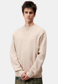 Sudadera beige de manga larga con puños acanalados, que presenta un pequeño logo blanco en el pecho, llevada sobre pantalones de camuflaje.