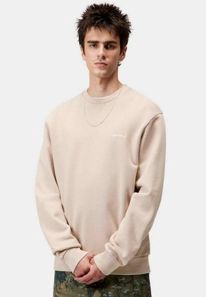 Sudadera beige de manga larga con puños acanalados, que presenta un pequeño logo blanco en el pecho, llevada sobre pantalones de camuflaje.