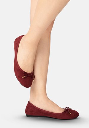 Ballerines bordeaux à talon bas avec petits détails de nœuds, portées sur des jambes croisées nues sur un fond uni.