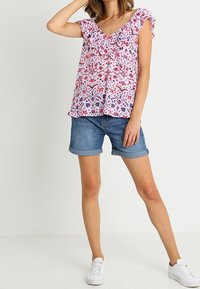 Femme portant un chemisier à manches courtes à fleurs roses, un short en jean retroussé et des baskets blanches, debout avec un bras levé et les jambes légèrement écartées.