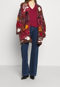 Cardigan en maille multicolore à motif floral, superposé à un pull bordeaux à col en V, associé à un jean évasé bleu et des bottes à talons noires.