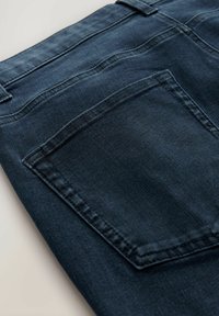 Tmavé denim džínsy majú zadnú kapsu s viditeľným stehom, hladkú textúru a zasúvajúci dizajn pásu. Farba je tmavomodrá.