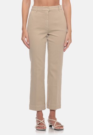 Donna che indossa pantaloni beige a gamba dritta e tacchi bianchi aperti con cinturini, in piedi davanti a uno sfondo semplice.
