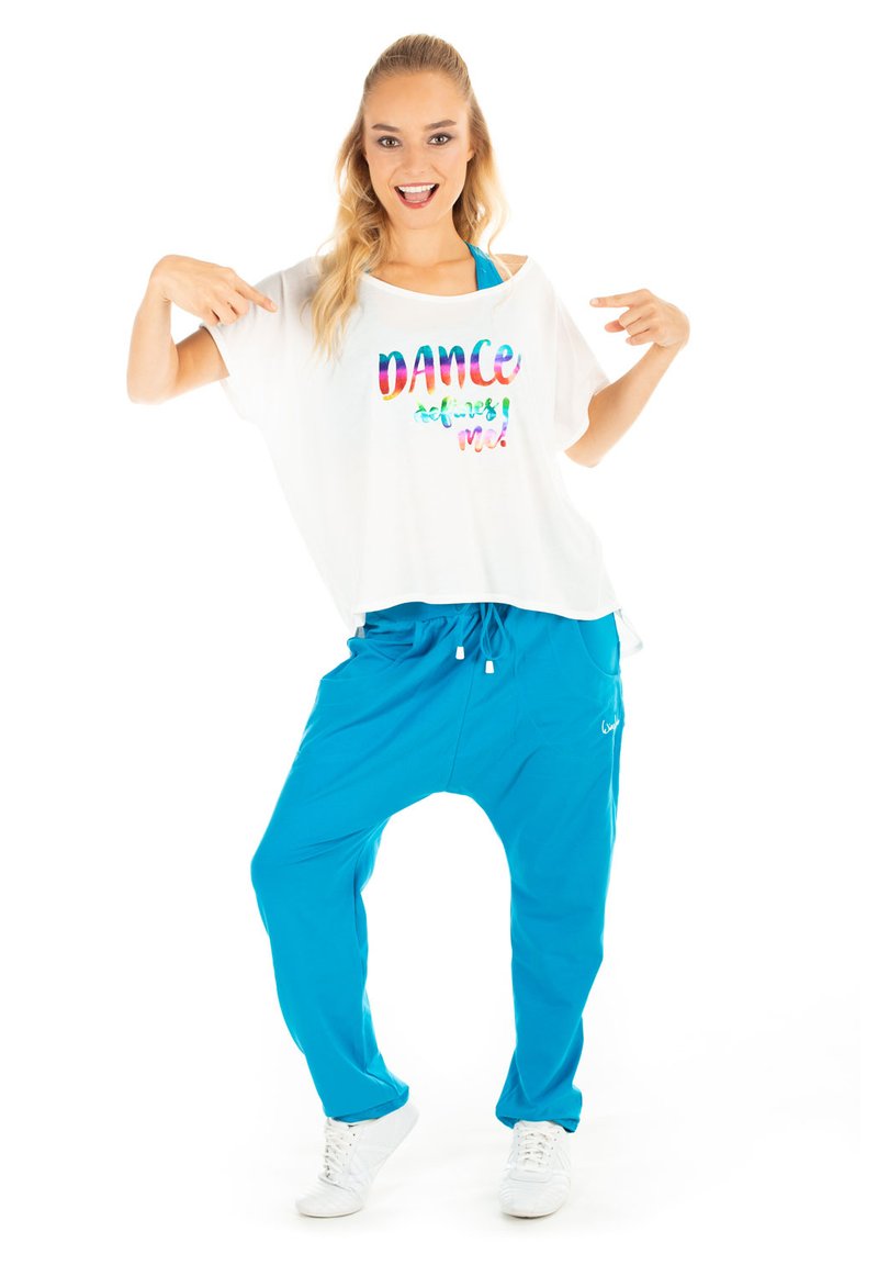 White oversized shirt with colorful "DANCE defines me!" text, paired with loose blue pants. Model wearing white sneakers, posing playfully.