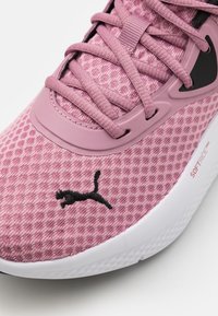 Różowy but sportowy z teksturowaną, siateczkową cholewką, z czarnymi akcentami i logo. Białą, wyściełaną podeszwę dopełnia całość.