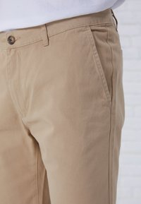 Beige cotton chinos with a smooth texture, featuring a side pocket and a button closure at the waistband. Close-up view highlights stitching.
