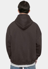 Dark brown hoodie with a relaxed fit, large hood, and ribbed hem. Worn over a white shirt, textured fabric visible on the surface.