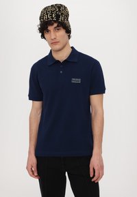 Mornarska polo majica s kratkimi rokavi, z gumbi spredaj in majhnim žepnim nalepkom na levem prsnem delu. Nosijo jo s teksturirano črno in zlatno kapo.