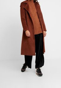 Cappotto lungo marrone con colletto, indossato sopra un maglione arancione a costine. Pantaloni neri plissettati e sneakers multicolori completano l'outfit.