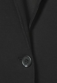 Zwarte stoffen blazer met een opstaande kraag en een enkele zwarte knoop met vier gaten, gepresenteerd tegen een gladde achtergrond.