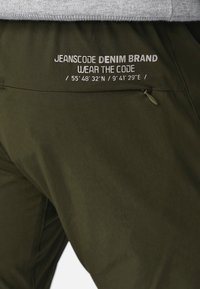 Olivové nohavice so zipsovým vreckom, s bielym textom, ktorý znie "JEANSCODE DENIM BRAND WEAR THE CODE" a geografickými súradnicami.