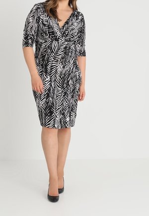 Femme portant une robe portefeuille noire et blanche à motif de feuilles, longueur genou, manches trois-quarts, et des talons noirs, sur un fond uni.