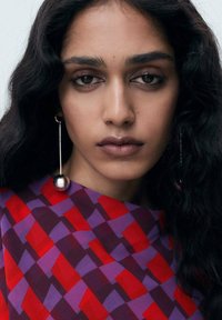 Red and purple patterned blouse with geometric shapes, featuring a silver spherical earring and long, wavy black hair.