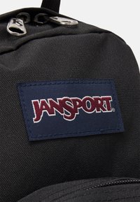Crni ruksak s teksturiranim materijalom, s plavom i bijelom oznakom Jansport i srebrnim patentnim zatvaračima na vrhu.